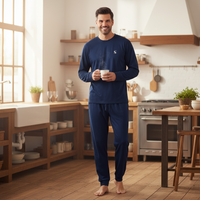 A man stands barefoot in a bright, modern kitchen wearing a navy blue long-sleeve pajama shirt and matching pants, holding a mug of coffee.
