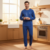 A man stands barefoot in a bright kitchen wearing a matching royal blue long-sleeve pajama set and holding a steaming mug. The kitchen features light wood cabinets, a white farmhouse sink, and a stainless steel oven.