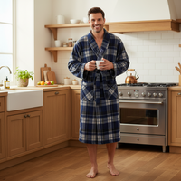 A man wearing a blue and white plaid bathrobe stands barefoot in a bright, modern kitchen, holding a white mug.