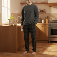 A man wearing a dark gray long-sleeve crewneck shirt and dark gray plaid pajama pants stands barefoot in a warm, sunlit kitchen.