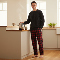 A man wearing a black long-sleeve shirt and red and black plaid pajama pants stands barefoot in a bright, modern kitchen, holding a silver kettle with steam rising.