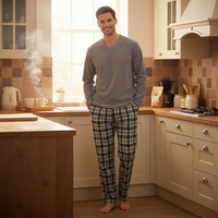 A man stands barefoot in a warm, sunlit kitchen wearing a gray long-sleeve V-neck shirt and green-and-white plaid pajama pants.