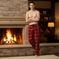 A shirtless man stands barefoot in front of a stone fireplace wearing red plaid pajama pants with black and white lines. The background shows a cozy living room with a lit fireplace, bookshelves, and a lamp.