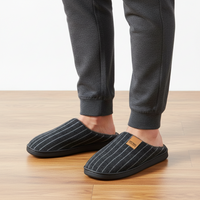 A person wearing dark gray pinstriped slip-on slippers with a small tan brand patch, paired with dark gray jogger pants, standing on a light wood floor against a white wall.