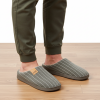 A person wearing olive green pants and gray pinstriped slippers with a soft fleece lining, standing on a light wood floor against a white background.