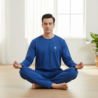 A man meditating in a bright, modern room, wearing a matching royal blue long-sleeve shirt and pants set with a small white embroidered logo on the chest.