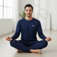 A man sits cross-legged in a meditative pose wearing a navy blue long-sleeve crewneck shirt and matching pants with a small white embroidered letter 'S' on the chest, in a bright, modern room with a white brick wall and light wood floor.
