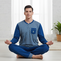 A man sits cross-legged in a meditative pose wearing a two-piece pajama set. The set features a long-sleeve henley top with blue and navy horizontal stripes on the front, solid blue sleeves, and a matching blue chest pocket with a white monogram 'S'.