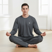A man sits cross-legged in a meditative pose wearing a dark gray long-sleeve shirt and matching pants in a bright, modern room with light wood floors and white walls.