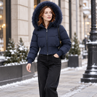 A woman walking outdoors in a snowy city setting wearing a dark navy puffer jacket with a large dark blue fur-trimmed hood, paired with black jeans and carrying a black shoulder bag.