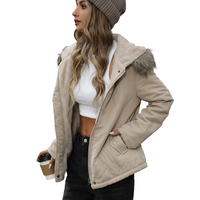 A woman wearing a beige hooded puffer jacket with a faux-fur trimmed hood, a white cropped top, black pants, and a gray beanie, holding a takeaway coffee cup against a plain white background.