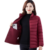 A woman wearing a burgundy puffer jacket over a black turtleneck sweater, holding open the jacket to show the interior pocket. The background is solid white.