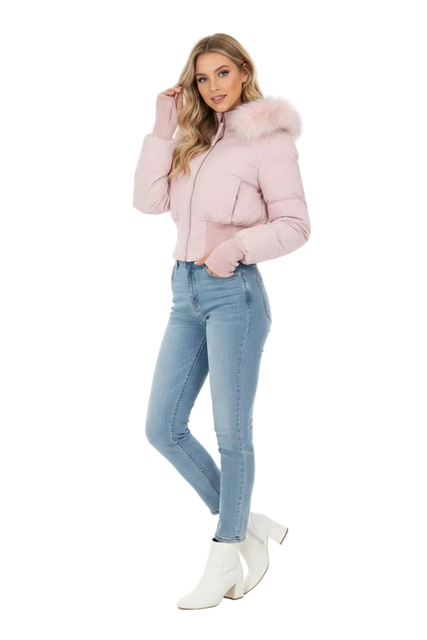 A woman modeling a light pink cropped puffer jacket with a fluffy faux-fur trimmed hood, paired with light-wash blue skinny jeans and white ankle boots, against a plain white studio background.