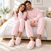 A smiling couple wearing matching light pink plush pajama sets and slip-on slippers sits together on a bed in a bright, modern bedroom.