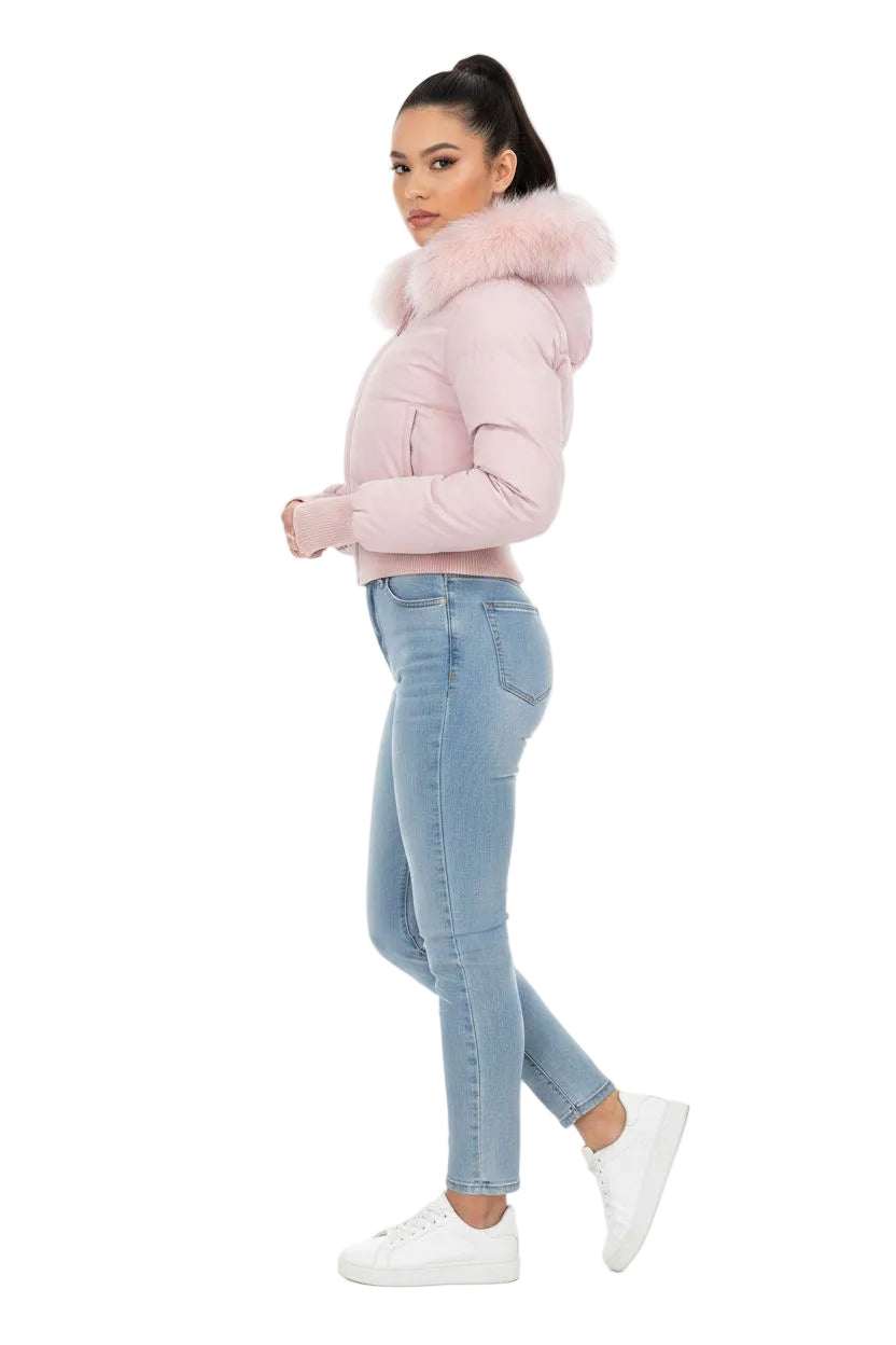 A woman modeling a light pink puffer jacket with a fluffy faux-fur trimmed hood, paired with light blue skinny jeans and white sneakers, against a plain white background.