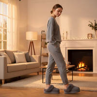 A woman stands in profile in a cozy living room wearing a matching gray knit lounge set with gray fuzzy slip-on slippers. The room features a fireplace, a beige sofa, and warm ambient lighting.