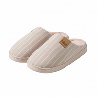 A pair of slip-on house slippers in a light beige color with thin white vertical stripes, featuring a small brown leather brand patch that reads 'HOME'.