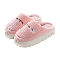 A pair of pink ribbed fleece slip-on slippers with white soles. One slipper features the text 'HAPPYLIFE' in black, and the other has a small embroidered bunny face.