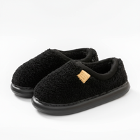 A pair of black fuzzy slip-on slippers with a small gold brand tag on the side, photographed on a clean white background.