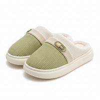 A pair of slip-on house slippers with a textured sage green upper and a cream ribbed knit collar, featuring a small green brand patch on the side and a thick cream sole.