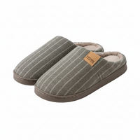 A pair of slip-on house slippers in a gray fabric with thin white vertical stripes, featuring a tan leather 'HOME' brand patch on the side and a soft beige lining, shown against a plain white background.