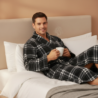 A man wearing a black and white plaid flannel robe and matching pants sits on a bed with white pillows and bedding, holding a white mug.