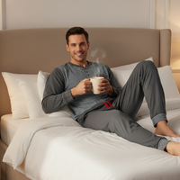 A man reclining on a bed wearing a gray long-sleeve striped pajama set and holding a steaming mug. The bedroom has a beige upholstered headboard, white bedding, and soft warm lighting.
