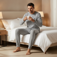 A man wearing a light gray long-sleeve pajama set sits on a bed with white linens, holding a white mug in a warmly lit bedroom.
