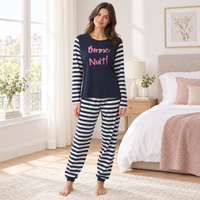 A woman stands barefoot in a bright bedroom wearing a navy and navy-and-white striped pajama set with pink 'Bonne Nuit!' text on the top. The room includes a bed, window, and bedside table.