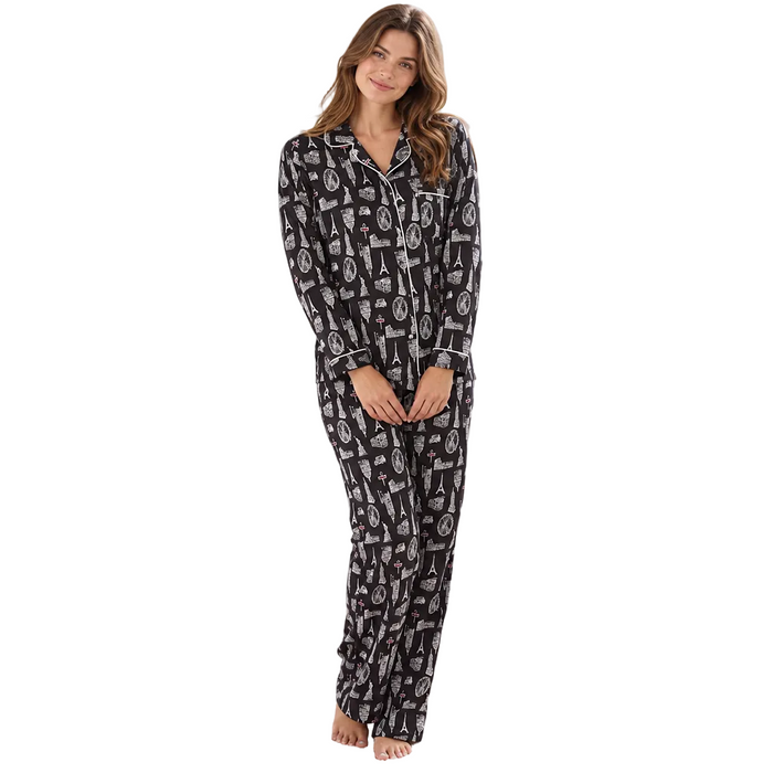 A woman stands barefoot in a bright bedroom wearing a black long-sleeve pajama set with a white illustrated pattern. The room includes a bed, white nightstand, lamp, window with daylight, and a framed picture on the wall.