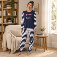 A woman stands barefoot in a cozy living room wearing a navy blue and white striped pajama set with 'Bonne Nuit!' printed in pink on the chest. The room features a wooden bookshelf, a cream armchair with a knitted throw, and a small wooden side table.