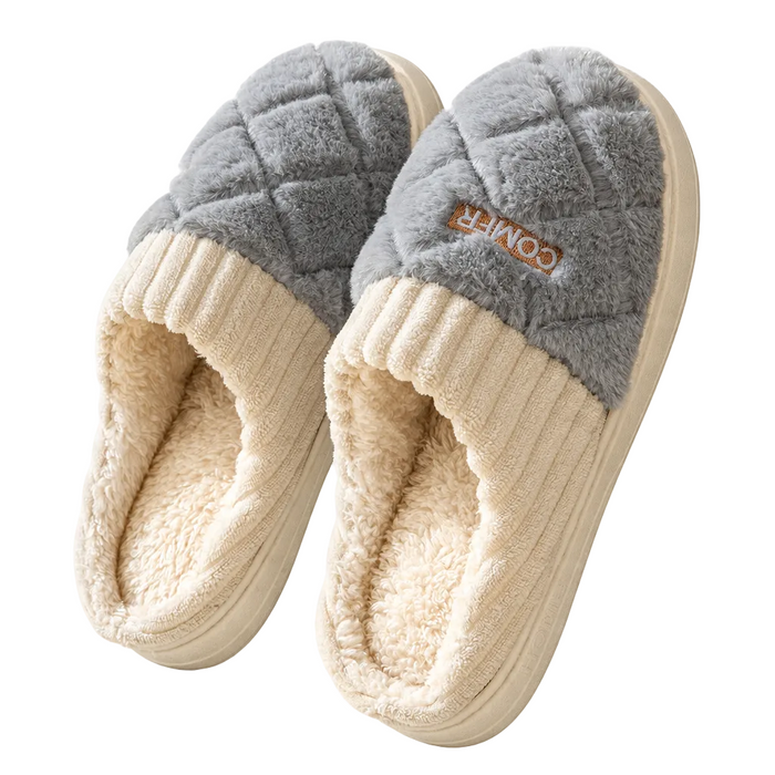 A pair of plush indoor slippers with a gray quilted upper and cream ribbed trim and lining, shown on a clean white background. A small brown brand patch on the side reads COMFY.