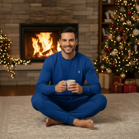 A man wearing a matching royal blue long-sleeve pajama set sits cross-legged on a patterned rug in front of a lit fireplace, holding a mug. A decorated Christmas tree and warm holiday lights are visible in the background.