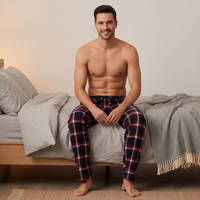 A shirtless man sits on a bed wearing navy plaid pajama pants with red and white lines, in a softly lit bedroom setting.