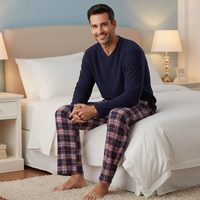 A man sits on a neatly made bed wearing a navy long-sleeve V-neck top and matching navy-and-red plaid pajama pants in a bright, modern bedroom.