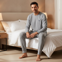 A man sits barefoot on a neatly made bed wearing a light gray long-sleeve pajama top and matching gray jogger pants in a warm, modern bedroom setting.