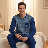 A man wearing a blue and navy striped long-sleeve pajama shirt with solid blue sleeves and a chest pocket, sitting on a bed in a softly lit bedroom.
