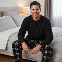 A smiling man sits on a bed in a softly lit bedroom, wearing a black long-sleeve crewneck shirt and plaid pajama pants in red, black, white, and gray.