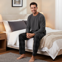 A man wearing a dark gray long-sleeve top and dark gray plaid pajama pants sits on a neatly made bed in a warm, modern bedroom.