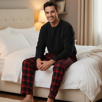 A man sitting on a bed wearing a black long-sleeve shirt and red and black plaid pajama pants in a cozy bedroom setting.