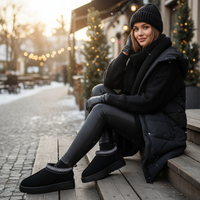 A woman sits on wooden steps outdoors in a snowy setting, wearing a black knit beanie, black knit sweater, black puffer vest, black leggings, and black slip-on winter boots with a patterned trim.