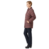 A woman stands in profile wearing a patterned quilted jacket with dark navy trousers and black ankle boots, photographed against a clean white studio background.