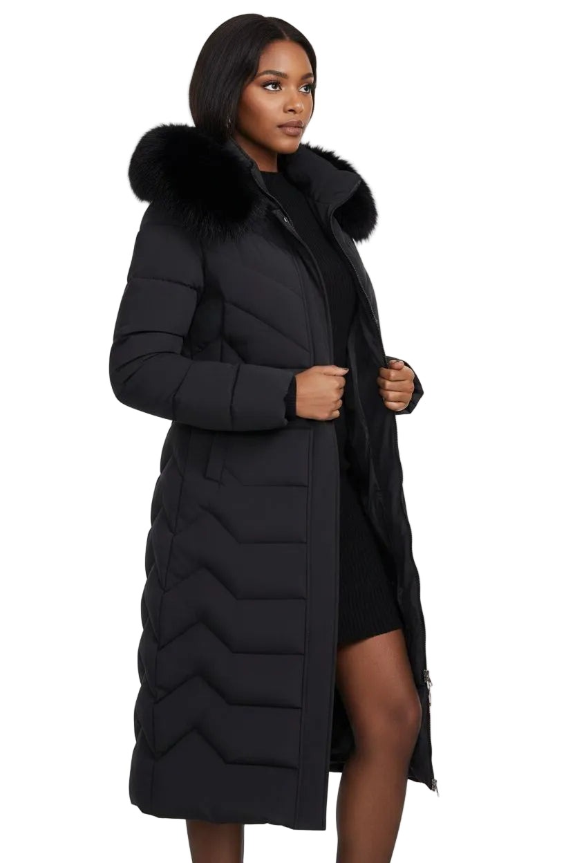 A woman modeling a long black quilted puffer coat with a large black faux-fur collar, photographed against a plain white studio background.