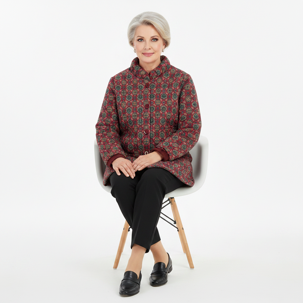 An older woman with short gray hair sits on a white chair with wooden legs, wearing a patterned quilted jacket in dark red, green, and gold, black pants, and black loafers against a plain white studio background.