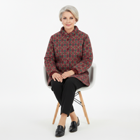 An older woman with short gray hair sits on a white chair with wooden legs, wearing a patterned quilted jacket in dark red, green, and gold, black pants, and black loafers against a plain white studio background.