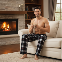 A shirtless man sitting on a cream sofa in a cozy living room, holding a mug. He is wearing dark plaid pajama pants with red, black, and white tones.