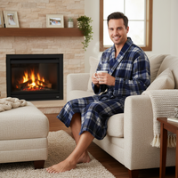A man wearing a blue and navy plaid bathrobe sits comfortably on a cream-colored armchair in a cozy living room with a lit fireplace, holding a white mug.