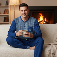 A man wearing a blue and navy striped long-sleeve pajama set sits cross-legged on a cream-colored couch, holding a ceramic mug in front of a warm fireplace and bookshelves.