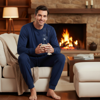 A man wearing a navy blue long-sleeve pajama set sits barefoot on a cream couch in a cozy living room with a lit fireplace and a mug in his hands.