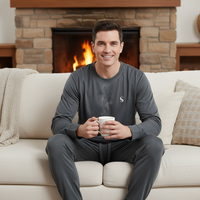 A man sits on a cream couch in front of a stone fireplace, wearing a dark gray long-sleeve crewneck shirt and matching gray jogger pants while holding a white mug.